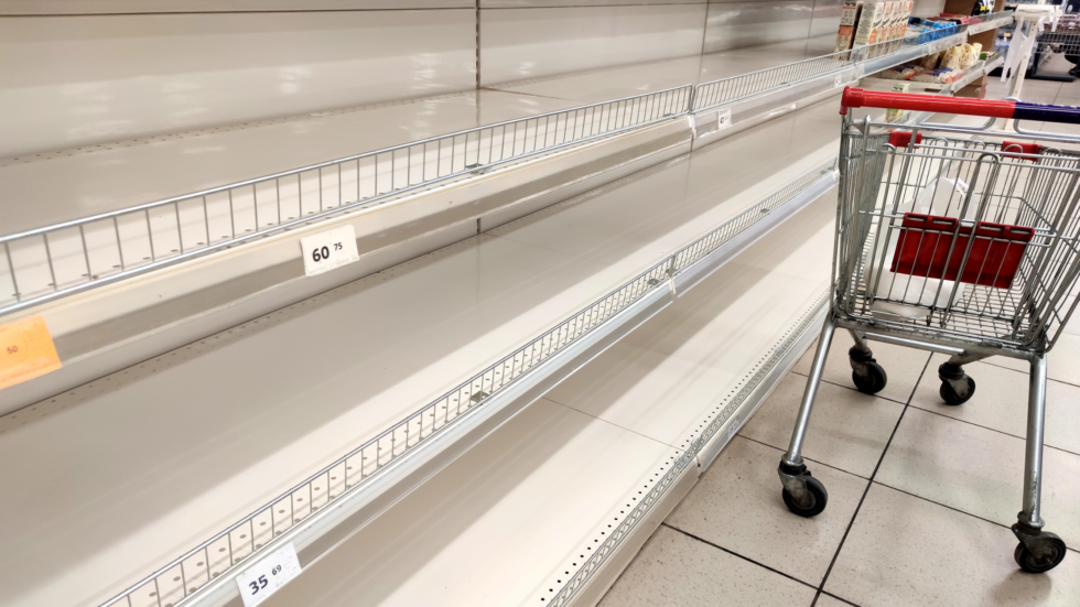 An empty metal grocery cart with a red handlebar next to empty shelves with price stickers in a store.