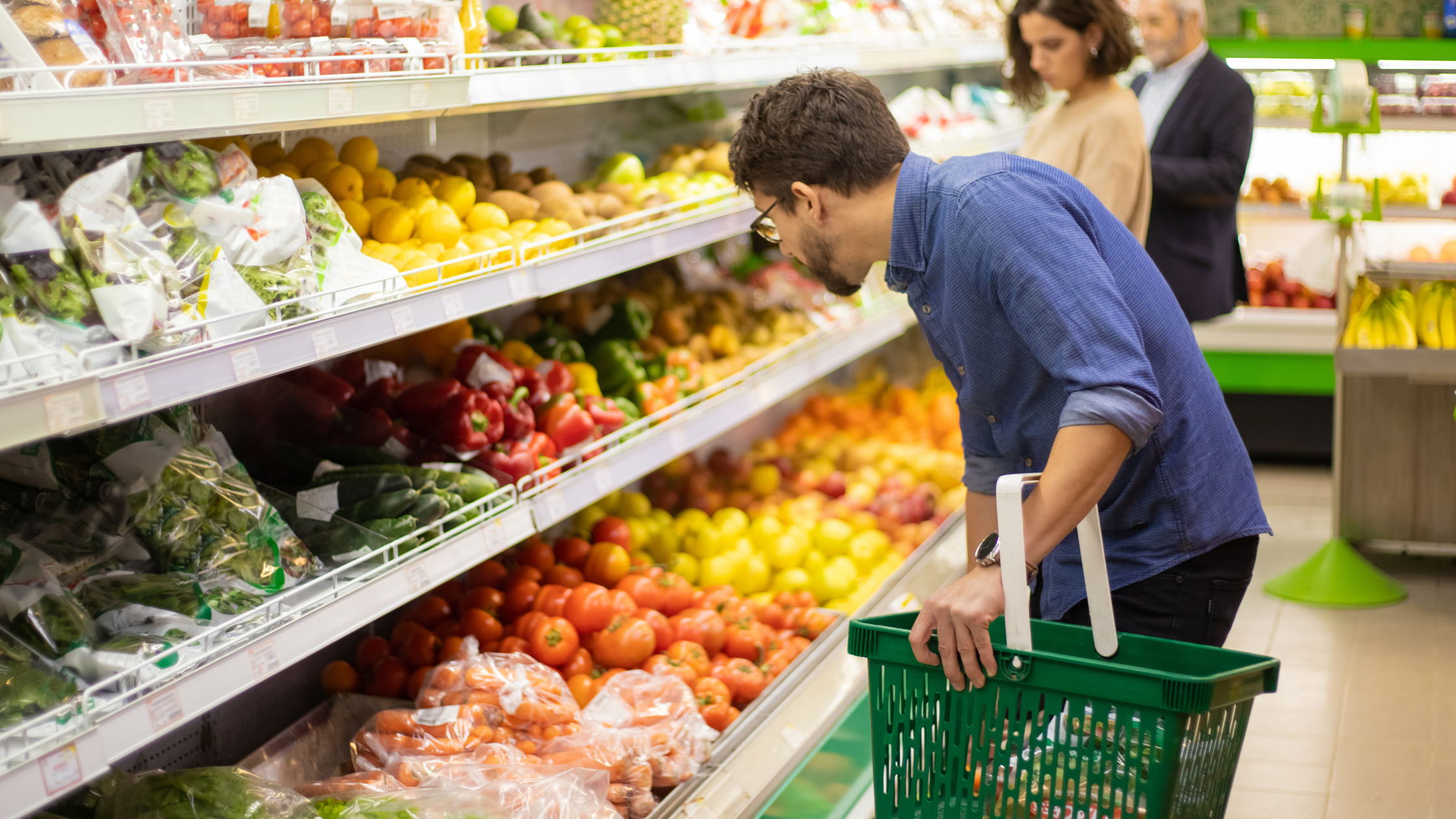 A brown-haired man with glasses and a beard holds a green grocery basket and looks at the organized produce section of a store