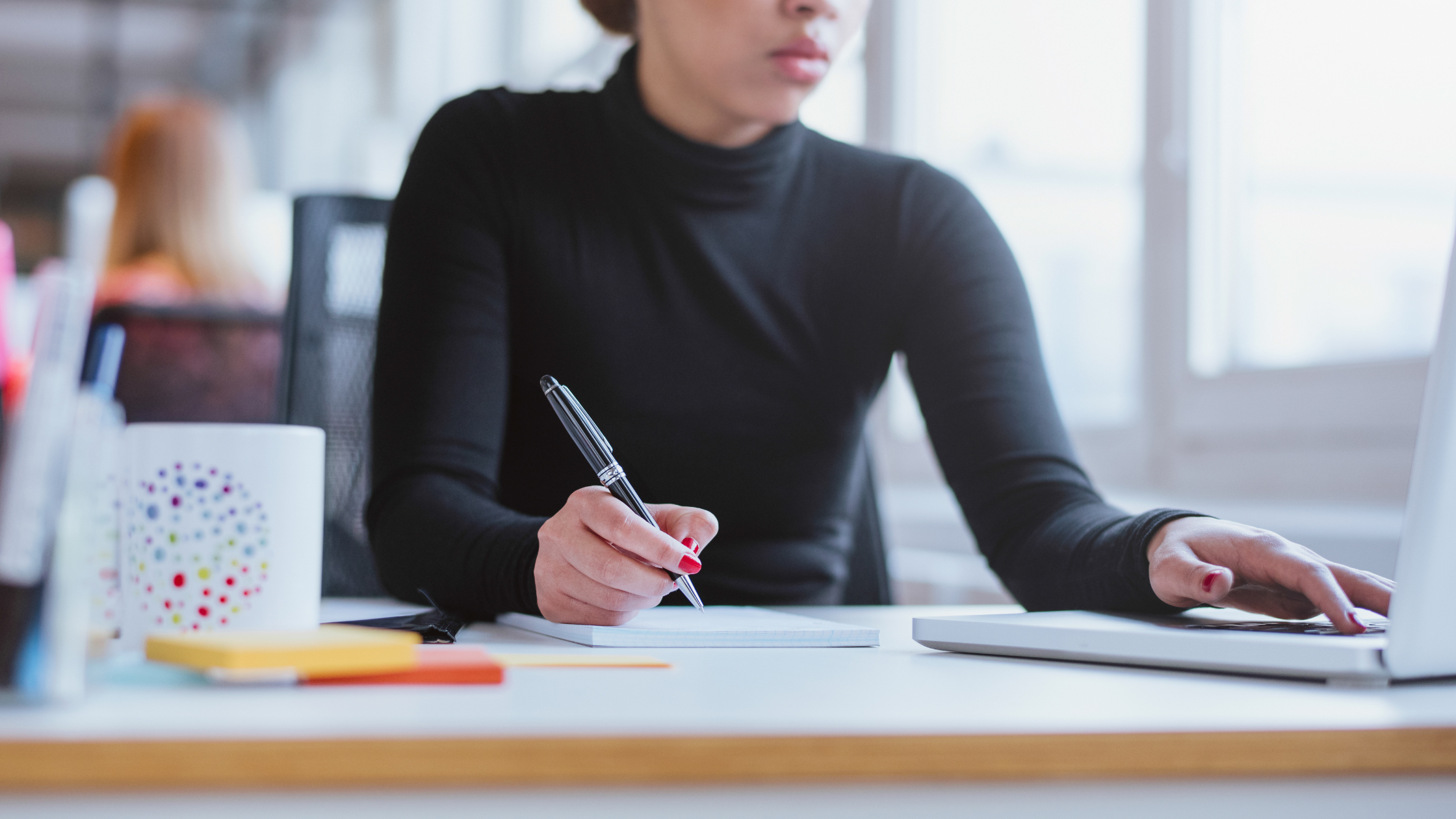 A woman in a black long-sleeve turtleneck sits at a desk with one hand scrolling on a laptop and the other writing in a notebook