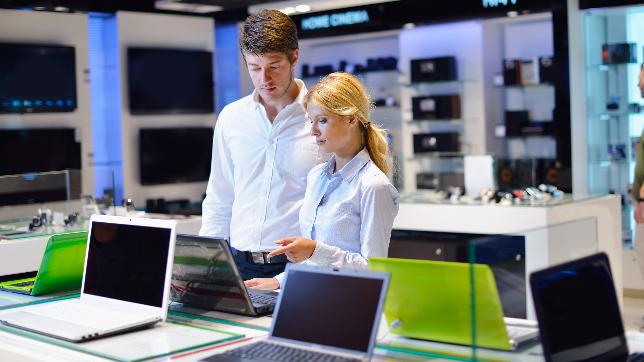 A blonde-haired woman and a tall man are looking at laptops in a store